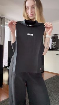 Junge Frau, die ein schwarzes ärmelloses Levi's Top hält, steht in einem Wohnzimmer, während ihr tätowierter Unterarm und eine Armbanduhr sichtbar sind.