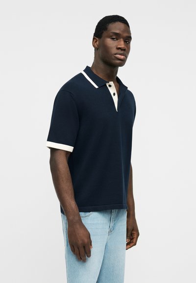 Άνδρας που φοράει κοντομάνικο μπλε μπλουζάκι πόλο με λευκό γιακά και άκρες μανικιών, σε συνδυασμό με ανοιχτόχρωμο τζιν, στέκεται μπροστά σε λευκό φόντο.