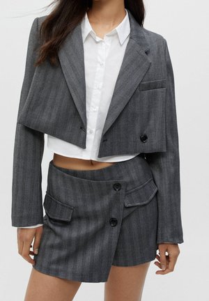 Frau trägt ein graues, kurzes Blazer und einen asymmetrisch geknöpften Rock dazu, über einer weißen, kurzen Hemdbluse mit Knöpfen.