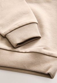 Beige Sweatshirt-Detail mit gerippten Bündchen und Saum, glatter Textur und weichem Stoff. Die Bündchen sind umgeschlagen und eng anliegend.