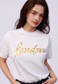 Camiseta de algodón blanca con un gráfico en cursiva dorada que dice "London". Manga corta y escote redondo con un corte relajado.