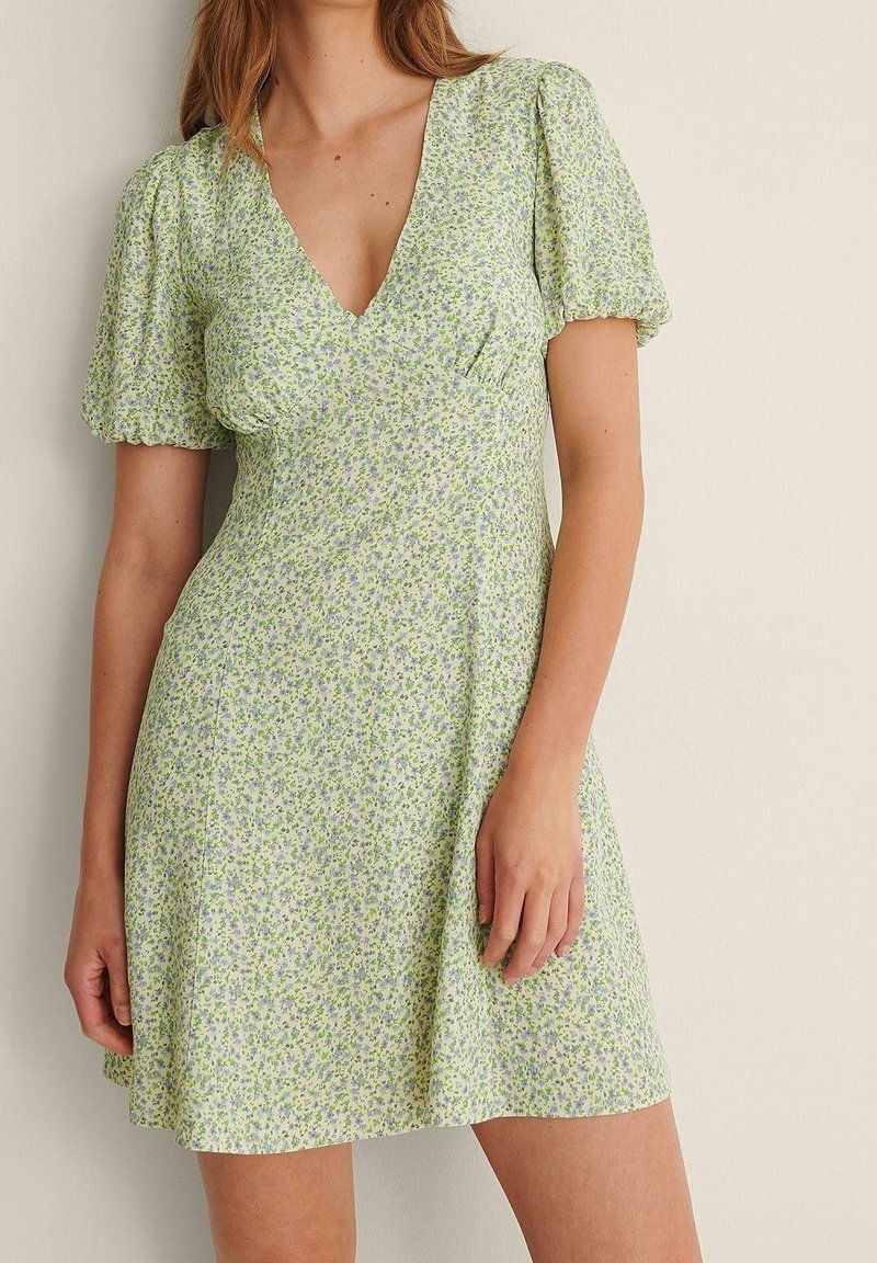 Mujer con un vestido corto de color verde claro con pequeño estampado floral azul y mangas cortas abullonadas, de pie contra una pared lisa.