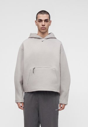 Giovane uomo con capelli corti che indossa una felpa con cappuccio oversize grigio chiaro e pantaloni della tuta grigio scuro su sfondo bianco semplice.