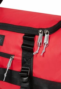 Sac en tissu rouge avec fermeture éclair et sangles noires, présentant des accessoires en métal argenté, des tirettes grises et une surface texturée. Éléments de design robustes visibles.
