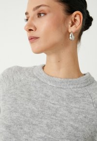 Grijze gebreide sweater met een ronde halslijn, gemaakt van een zachte, gestructureerde stof. Model draagt gepolijste zilveren oorhangers.