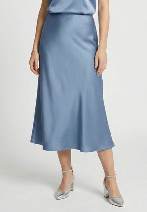Frau trägt einen blauen Satin-Midi-Rock kombiniert mit silbernen hochhackigen Knöchelriemchen-Schuhen, stehend vor schlichtem Hintergrund.