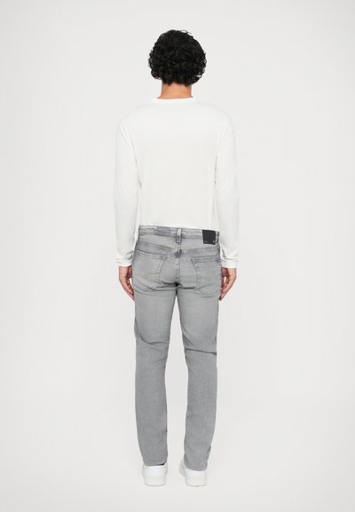 Leichte weiße Langarmbluse, graue Denim-Jeans mit einer aufgenähten Gesäßtasche und weiße Sneakers, von hinten betrachtet.