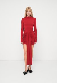 Piros, hosszú ujjú ruha, álló gallérral, ráncolt oldali részlettel, aszimmetrikus redőzéssel és sima textúrával. Fekete, magassarkú cipővel párosítva.