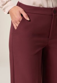 Pantaloni a gamba larga borgogna con una texture liscia, tasche laterali e chiusura a bottone in vita.