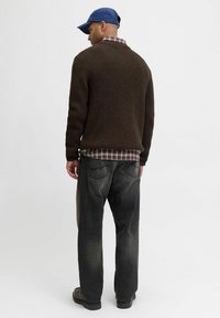 Maglione in tessuto a maglia marrone con texture a coste, indossato sopra una camicia a quadri rossa e bianca, abbinato a jeans scuri e un cappellino blu.