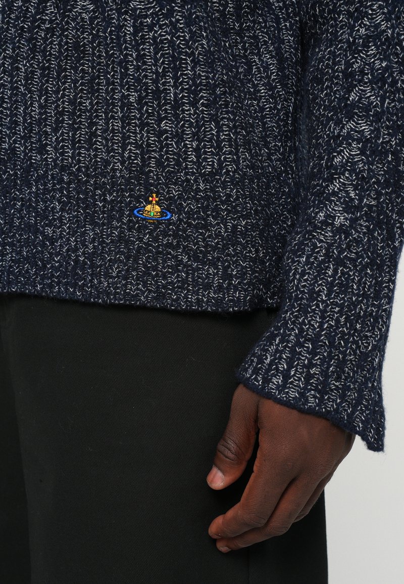 Nahaufnahme einer dunkelhäutigen Hand neben einem marineblauen und weißen Strickpullover mit einem kleinen bunten Orb-Logo, kombiniert mit schwarzen Hosen.