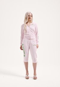 Rózsaszín cipzáras kapucnis pulóver és rövidnadrág puha anyagból, zöld felirattal. A nadrág alja állítható megkötőkkel, derékrésze pedig húzózsinóros.