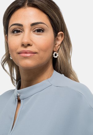 Blusa azzurro chiaro con un colletto alto e un bottone decorativo, abbinata a orecchini a cerchio in perle con un accento dorato.