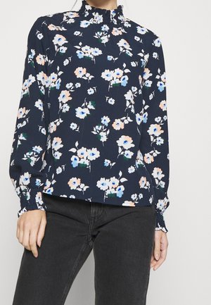 Mujer que lleva una blusa azul marino con estampado floral en azul y melocotón, mangas largas abullonadas y cuello alto con volantes, combinada con vaqueros negros.