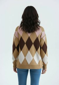 Kobieta o falowanych ciemnych włosach, ubrana w brązowy, różowy, biały i ciemnobrązowy sweter w romby oraz niebieskie dżinsy, stojąca tyłem na jednolitym tle.