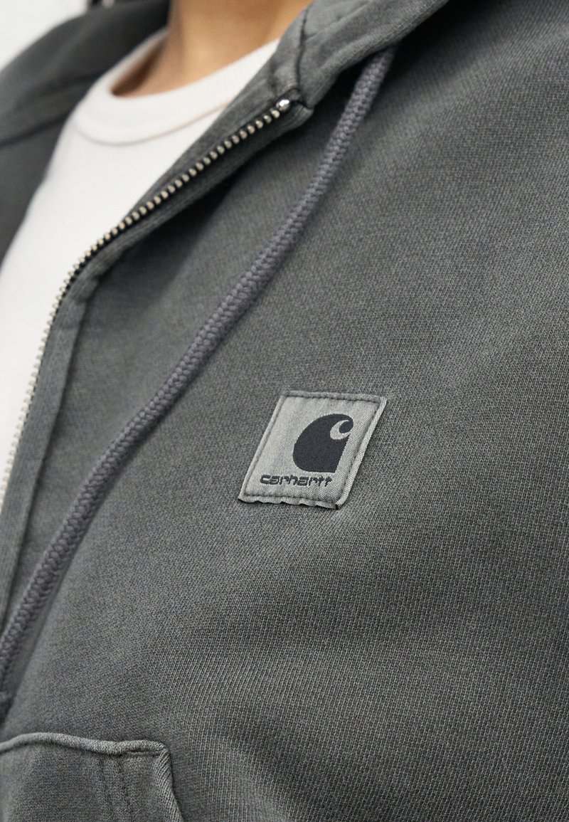 Primo piano di una persona che indossa una felpa con cappuccio Carhartt grigia con zip sopra una maglietta bianca, con il logo Carhartt cucito sul petto.