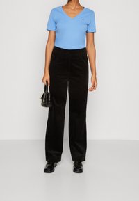 Světle modrý top s výstřihem do V a krátkým rukávem v kombinaci s černými kordovými širokými kalhotami, doplněný malým logem. Modelka drží černou kabelku.