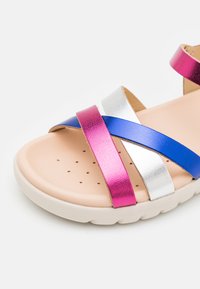 Sandale multicolore avec des brides métalliques roses, bleues et argentées. Semelle intérieure en beige texturé avec des trous de ventilation et une semelle en caoutchouc blanche.