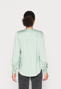 Blusa di seta di colore verde chiaro, con maniche lunghe e voluminose. Presenta un orlo curvo e pieghe sul retro. Scollo semplice con un piccolo dettaglio dell'etichetta.