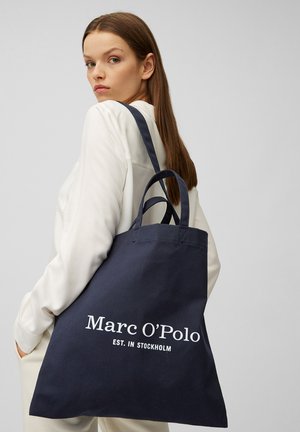 Frau mit langen braunen Haaren, die ein weißes Outfit trägt und eine große navyblaue Tragetasche mit dem Text "Marc O'Polo EST. IN STOCKHOLM" dabei hat.