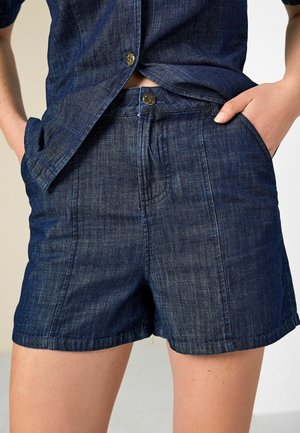 Dunkelblaue Denim-Shorts mit Fronttaschen, Knopf- und Reißverschluss, getragen mit einem passenden Denimhemd, das leicht in der Taille steckt.