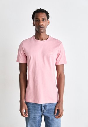 Camiseta de algodón color rosa claro, con mangas cortas, cuello redondo y un pequeño parche con logo en el pecho, combinada con jeans azules.