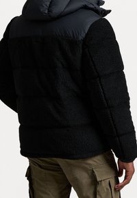 Person som bär en svart fleecetröja med en vadderad övre del och olivgröna cargobyxor, visad från baksidan och sidan.