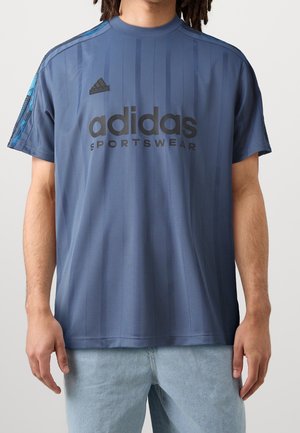 Mand iført blå Adidas sportstrøje og lyseblå jeans står mod en ensfarvet baggrund.