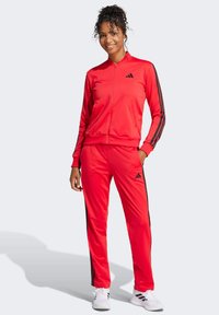 Rode sportieve trainingspak van gladde stof, met zwarte strepen aan de zijkanten en een adidas-logo op de jas en de broek.