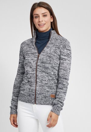 Grå og hvit teksturert zip-up genser med høy krage, med et brunt lærmerke og ribbede mansjetter. Buret over en marineblå rullekrage.