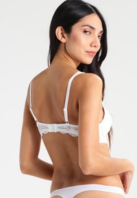 Soutien-gorge blanc à dos en dentelle avec bretelles ajustables, doté d'une fermeture à crochet et de délicats détails en dentelle sur les bords, en tissu lisse.
