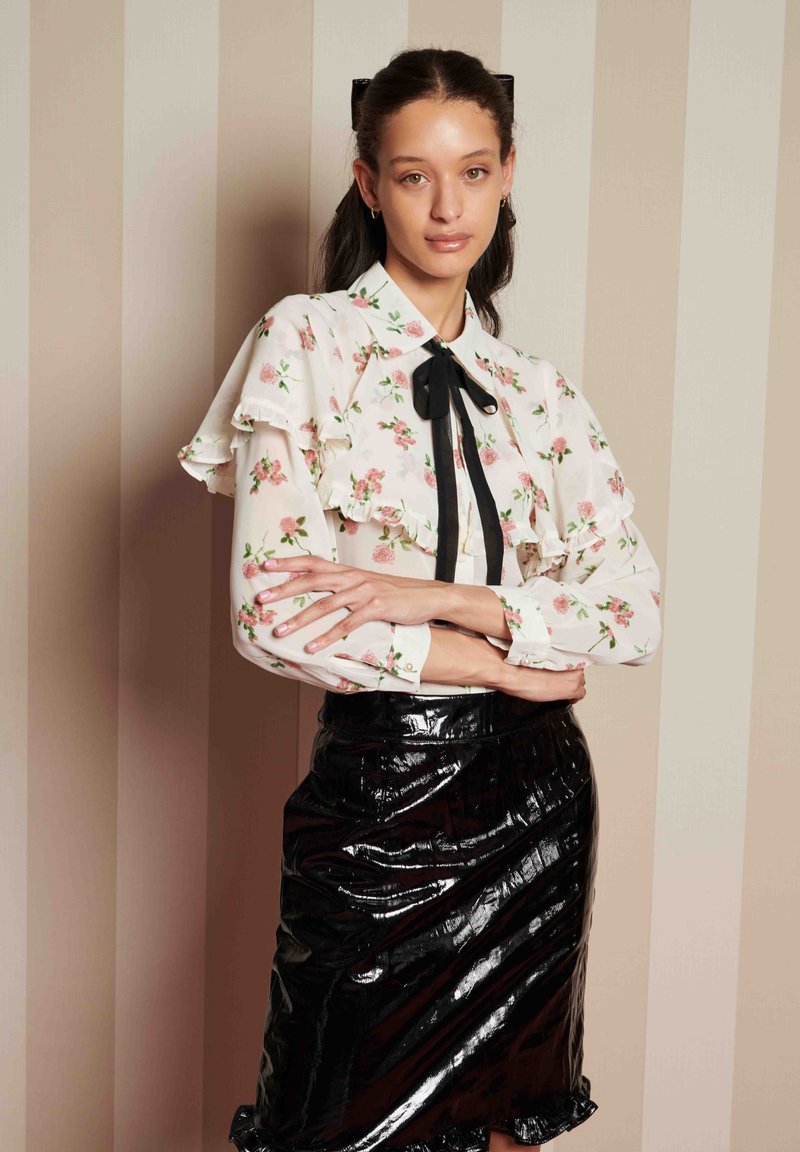 Blusa branca com padrão floral, mangas bufantes e laço preto, combinada com uma saia lápis preta brilhante com uma bainha rodada.