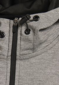 Haut à capuche zippé gris avec des accents noirs. Il dispose de cordons de serrage, d'une finition texturée et d'une fermeture éclair centrale. Le tissu semble doux et durable.