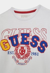 T-shirt in cotone biologico bianco con logo colorato "GUESS" in giallo, rosso e blu, decorato con un design di alloro e la scritta "EST. 1981".
