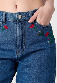 Džínové kalhoty mají modrou tkaninu s červenou květinovou výšivkou na předních kapsách, klasický design s pěti kapsami a knoflíkové zapínání v pase.