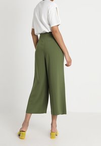 Henkilö, joka on pukeutunut korkeavyötäröisiin, leveälahkeisiin oliivinvihreisiin housuihin, valkoiseen lyhythihaiseen toppiin ja keltaisiin lohkokorkoisiin sandaaleihin, seisoo valkoista taustaa vasten.