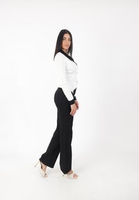 Cardigan côtelé blanc avec des accents noirs, associé à un pantalon noir évasé et des talons blancs à lanières. Le modèle se tient de profil sur fond blanc.