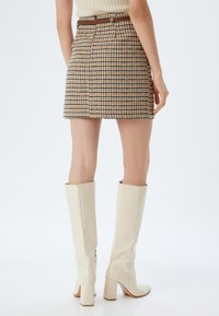 Braun-beige Hahnentrittmuster-Minirock mit Seitentaschen, Gürtelschlaufen und taillierter Silhouette, kombiniert mit hohen weißen Stiefeln mit Absätzen.