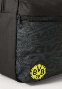 Schwarzer Rucksack aus strukturiertem Stoff mit einem Reißverschlussfach. Hat einen gelben runden Aufkleber mit "BVB 09" in fetten Buchstaben.