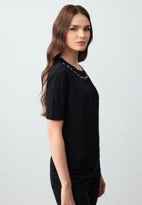 Zwarte trui met korte mouwen, gemaakt van een gladde stof en met een ronde halslijn, versierd met decoratieve accenten van rhinestones langs de kraag.