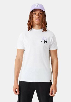 Giovane con lunghi dreadlocks che indossa una t-shirt bianca con un piccolo logo NY e un cappellino da baseball viola chiaro davanti a uno sfondo semplice.