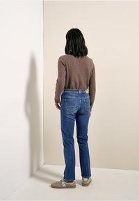 Vaqueros azules de corte recto, combinados con un suéter marrón texturizado y zapatillas deportivas grises. El fondo es neutro y liso.