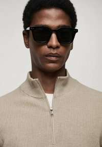 Beige ribbed zip-up sweater with a high collar, worn over a white T-shirt. Black round sunglasses add contrast to the neutral palette.