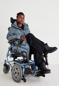 Elektrinė neįgaliųjų vežimėlis su šviesiai mėlynu rėmu, pilka pūkinė striukė, juodos briaunotos kelnės ir juodi tekstūruoti batai. Paprasta fono.