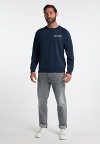 Granatowy sweatshirt z okrągłym dekoltem i rękawami raglanowymi, zestawiony z jasnoszarymi dżinsami i białymi sneakersami. Posiada nadrukowany tekst na klatce piersiowej.