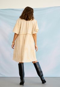 Femme aux cheveux bouclés portant une robe mi-longue brodée crème et des bottes noires à talons hauteur genou, debout devant un fond bleu clair.