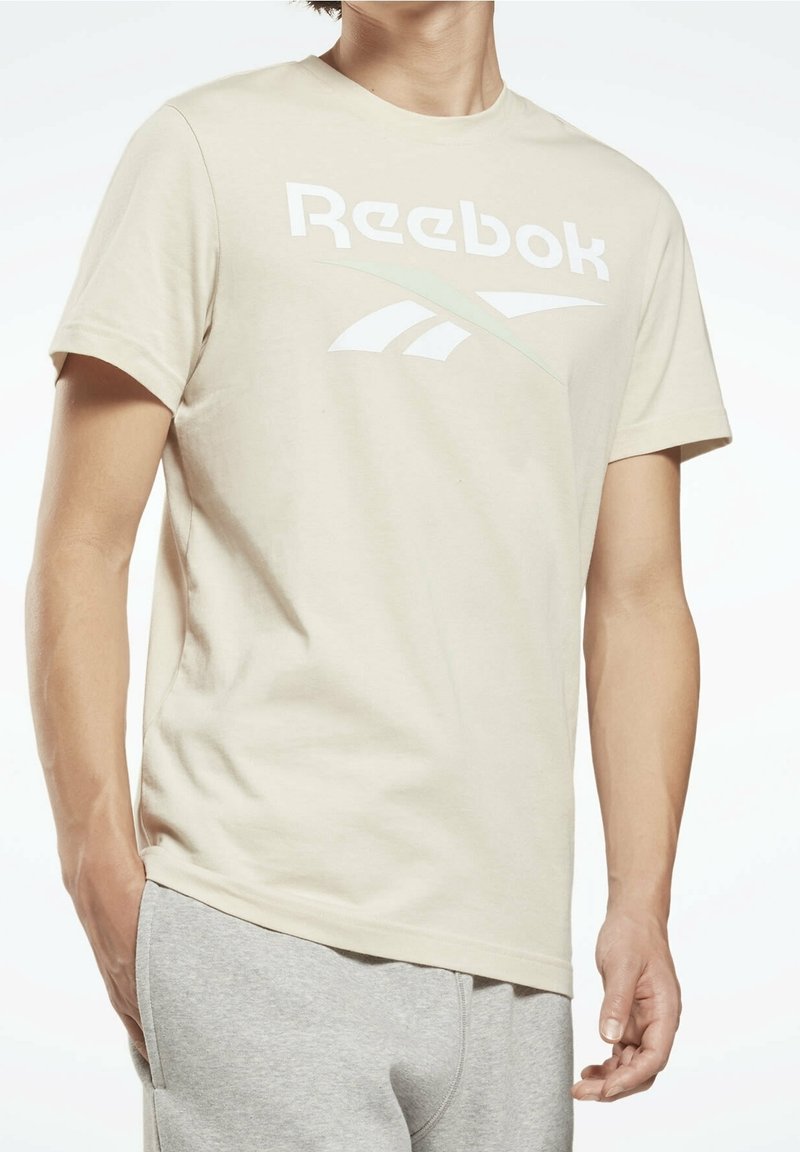 Beige bomullströja med en stor vit Reebok-logotyp och en subtil grön accent. Kort ärm och normal passform.