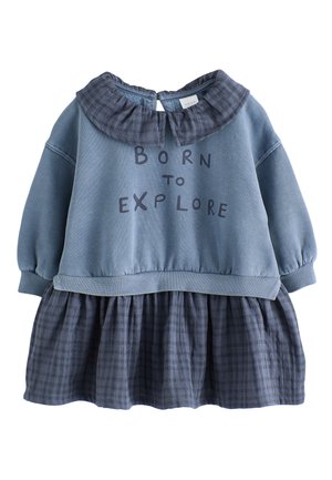 Mladé dievča s dvoma nadýchanými copmi, oblečené v modrých šatách s textom "Born to Explore", béžovými topánkami a tmavými ponožkami, stojí na béžovom pozadí.