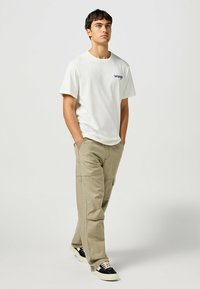 Camiseta de algodón blanca con logo azul en el pecho, combinada con pantalones cargo en color caqui claro que tienen múltiples bolsillos, completada con zapatillas negras y crema.
