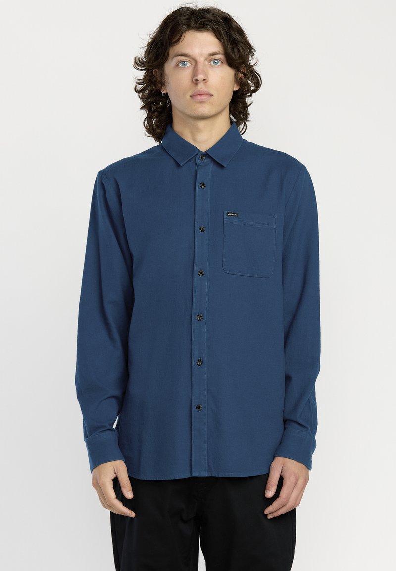 Chemise à manches longues en tissu bleu foncé, dotée d'une poche poitrine et de boutons noirs, avec un col et une coupe standard.